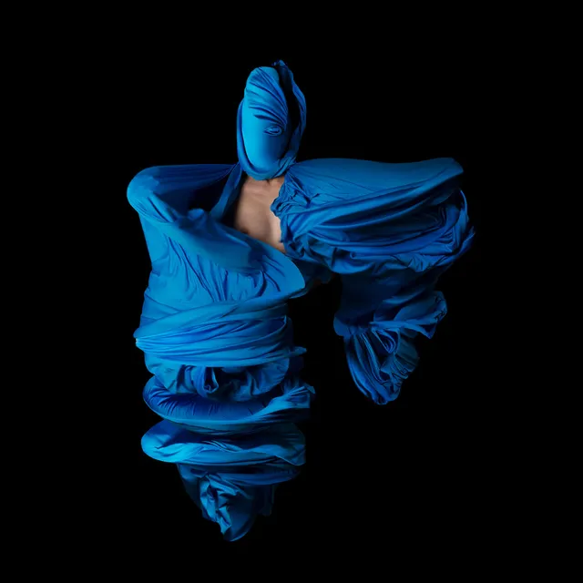 Fotografía performática de Lizette Abraham: figura envuelta en telas sobre fondo negro, con tela azul