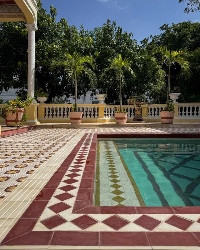 Alberca de azulejos rojos y verdes junto al corredor de columnas, con palmeras alrededor